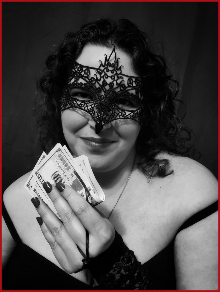 Mistriss Lilith is smiling patronizingly at the camera in this black and white photo. Her dark curly hair is down on her shoulders and she is wearing a black lace face mask over her eyes. In her hands she is holding a stack of $100 bills.